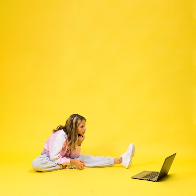 Il bambino positivo si siede con il computer portatile che legge i compiti indossando un panno in stile casual isolato su sfondo giallo