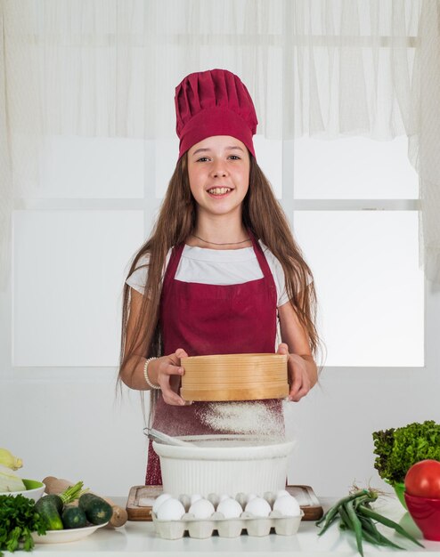 Il bambino prepara cibo sano a casa e indossa l'uniforme del cuoco per le pulizie e la casa aiutando lo sviluppo dell'infanzia piccola ragazza che cucina in cucina chef del bambino che cucina con la farina Cibo con il pensiero