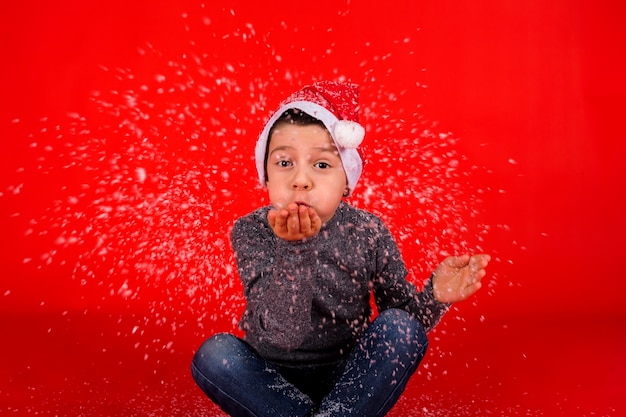 Il bambino ragazzo soffia la neve artificiale dalla sua mano su uno sfondo rosso