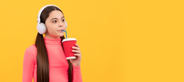 Il bambino serio ascolta la musica in cuffia beve dal bicchiere di carta su sfondo giallo caffè Poster orizzontale dello spazio di copia dell'intestazione del banner del viso del bambino isolato