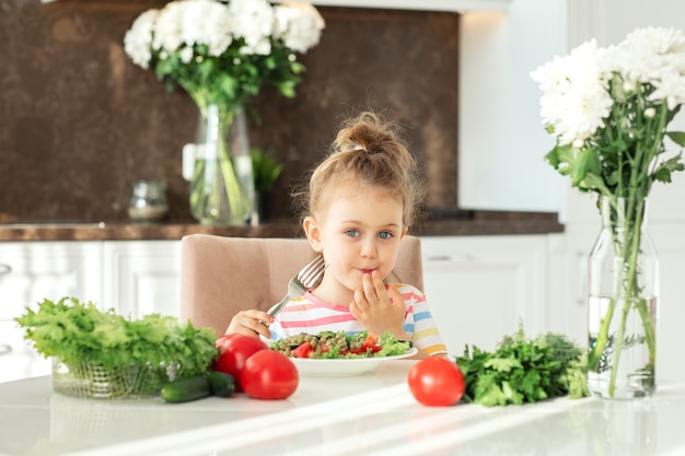 Il bambino si diverte a mangiare una sana insalata in cucina bianca soleggiata verdure fresche nutrizione concetto vegano