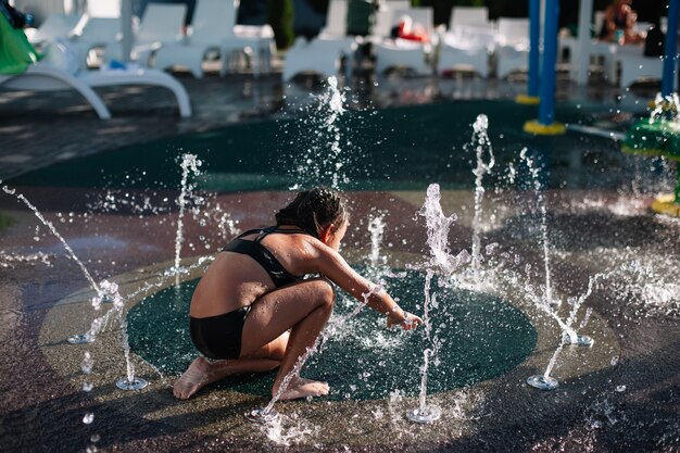 Il bambino si diverte all'attrazione dell'acqua La bambina in costume da bagno nero si è seduta e gioca con la fontana...