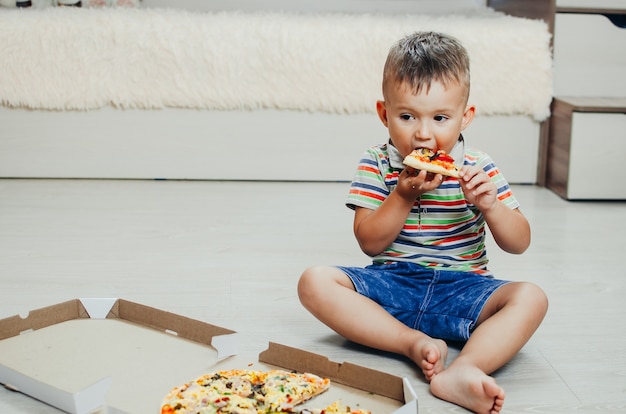 Il bambino si siede per terra e mangia la pizza molto appetitosa e golosa, in pantaloncini e maglietta