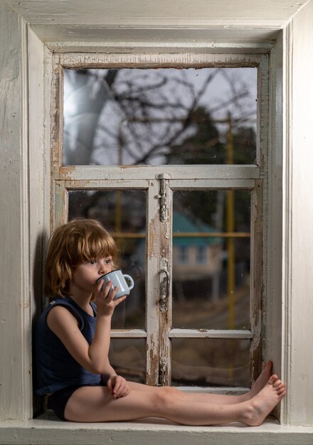 Il bambino si siede su un davanzale in legno squallido che beve da una tazza
