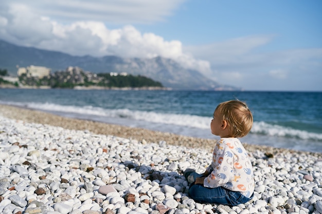 Il bambino si siede su una spiaggia di ciottoli in riva al mare