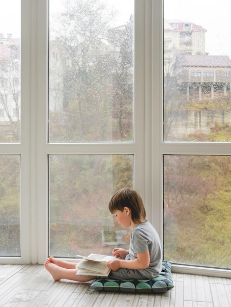 Il bambino si siede sul pavimento e legge un libro di svago intellettuale per bambini mentre fuori piove