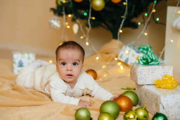 Il bambino sorridente sveglio è sdraiato sotto un albero di Natale festivo e gioca con i regali