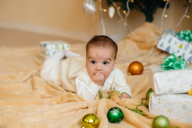 Il bambino sorridente sveglio è sdraiato sotto un albero di Natale festivo e gioca con i regali