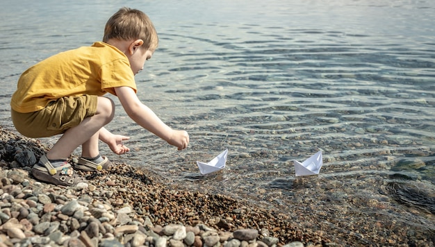 Il bambino sta lanciando barche di carta bianca nell'acqua limpida del mare
