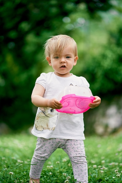 Il bambino sta su un prato verde con un contenitore di plastica per alimenti nelle sue mani