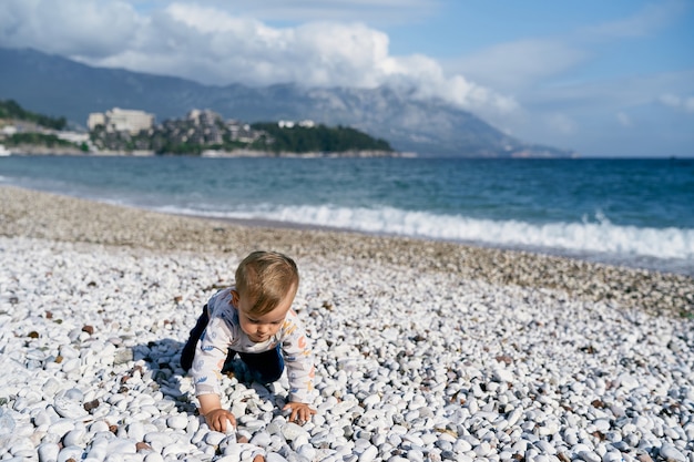 Il bambino striscia lungo la spiaggia di ciottoli sullo sfondo del mare e delle montagne