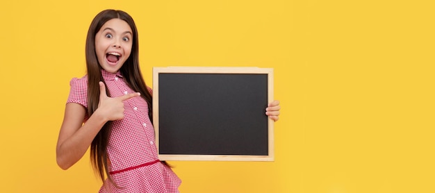 Il bambino stupito tiene la lavagna della scuola per copiare lo spazio che mostra il pollice in su Ritratto di intestazione del banner dello studio dello studente della studentessa Copyspace del viso del bambino della scuola