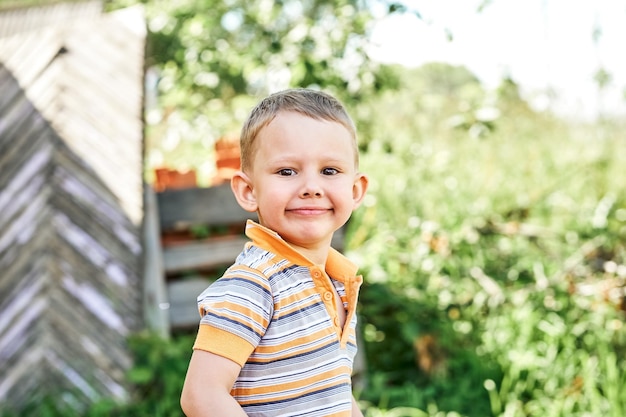 Il bambino sveglio con i capelli biondi sorride ampiamente in piedi nel cortile su sfondo sfocato