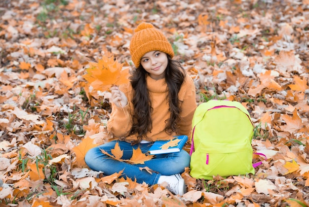 Il bambino sveglio in cappello giallo tiene la foglia d'acero all'aperto con lo zaino della scuola si rilassa nell'autunno del parco