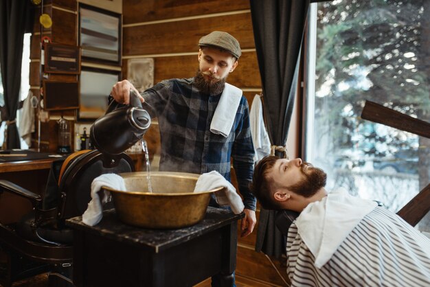 Il barbiere bagna l'asciugamano, taglia la barba. Il barbiere professionista è un'occupazione alla moda. Parrucchiere maschio e cliente nel parrucchiere