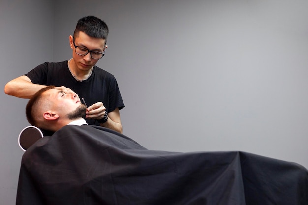 Il barbiere kazako si rade la barba con un rasoio a un cliente in un negozio di barbiere un ragazzo si sta facendo la barba