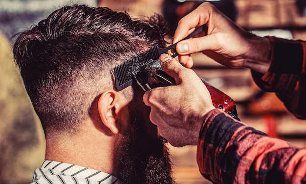 Il barbiere lavora con il tagliacapelli Cliente hipster che si taglia i capelli Mani del barbiere con il tagliacapelli da vicino Uomo barbuto dal barbiere Uomo che visita il parrucchiere dal barbiere
