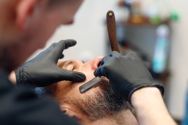 Il barbiere professionista rade la barba del cliente con un rasoio a mano libera. Barba tagliata con lama vecchio stile dal barbiere. Bell'uomo macho che si fa rasare la barba in studio. Colpo del primo piano.