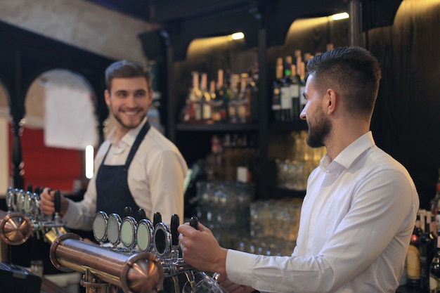 Il barista bello sorride e riempie un bicchiere di birra mentre si trova al bancone del bar nel pub.