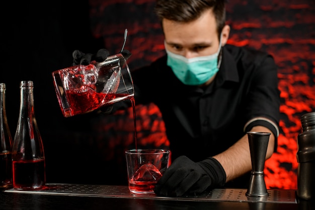 Il barista con la maschera versa con cura il cocktail freddo nel bicchiere