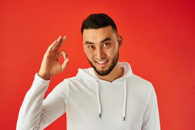 Il bel sorriso a trentadue denti dell'uomo kazako su sfondo rosso mostra il gesto "va bene". Ritratto di giovane ragazzo barbuto asiatico felice vestito di bianco casual isolato in studio