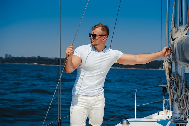 Il bellissimo giovane muscoloso in abiti bianchi si diverte a camminare sullo yacht mentre guarda il mare blu tu...