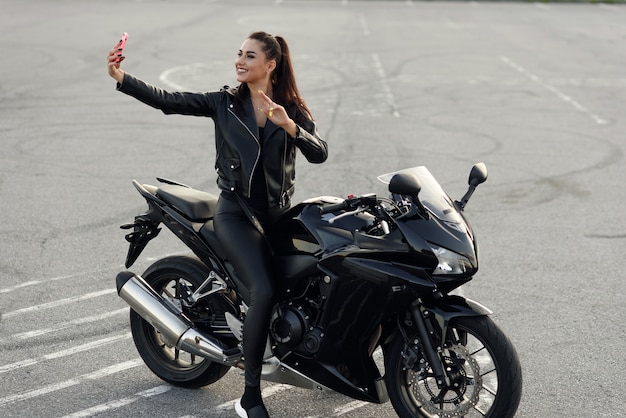 Il bello motociclista femminile fa la foto di selphie dallo smartphone mentre si siede sulla motocicletta sportiva alla moda