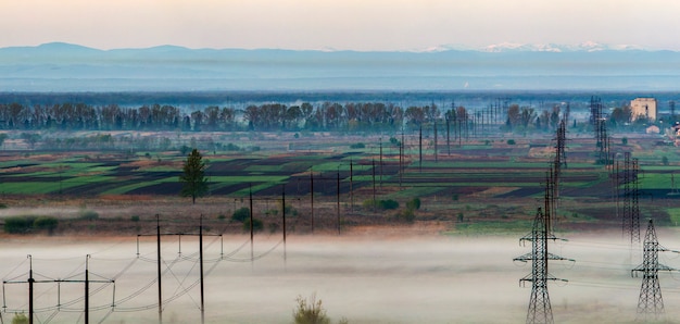 Il bello panorama della linea elettrica ad alta tensione elettrica lunga fila allungando all'orizzonte attraverso i campi e gli alberi della molla sotto la nebbia densa di mattina sul fondo distante della gamma di montagne carpatiche.