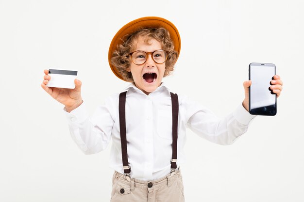 Il bello ragazzo con capelli ricci in camicia bianca, cappello marrone, vetri con le bretelle nere mostra un telefono e una carta dei soldi isolata su bianco