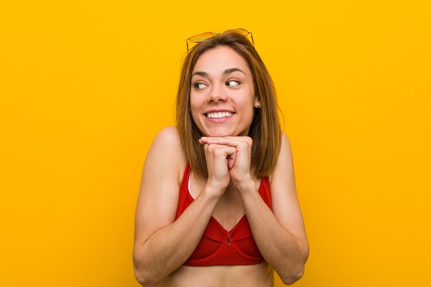 Il bikini e gli occhiali da sole da portare della giovane donna caucasica tengono le mani sotto il mento, stanno guardando felicemente da parte.