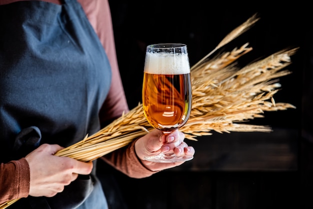 Il birraio in grembiule tiene un bicchiere di birra e spighette di grano