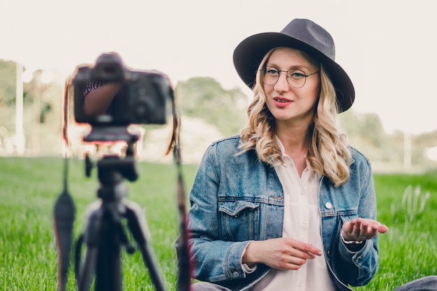 Il blogger alla moda della ragazza che si siede nel parco e spara il vlog sulla macchina fotografica.