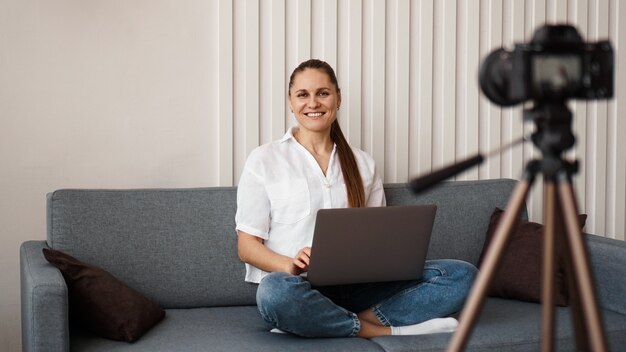 Il blogger femminile sorridente registra un nuovo video. Si siede sul divano a casa e tiene in mano un laptop. Concetto di blog aziendale positivo.