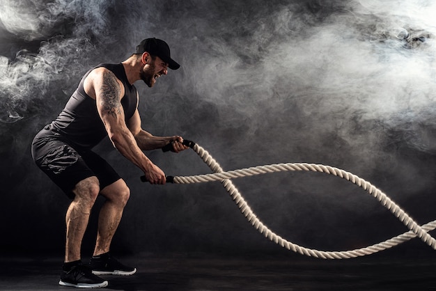 Il bodybulder dall'aspetto atletico barbuto si allena con la corda da battaglia sul buio con il fumo. Forza e motivazione.