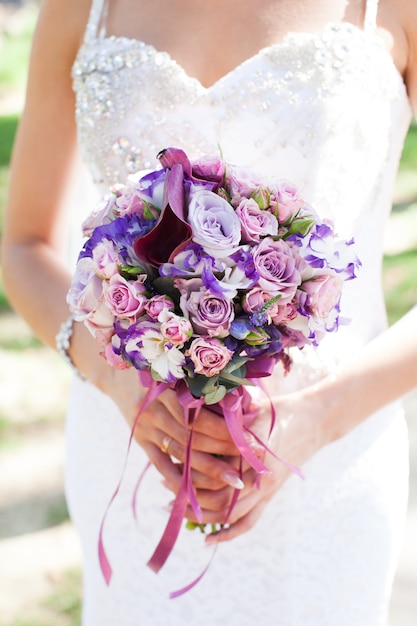 Il bouquet da sposa con rose lilla, eustoma e giglio nelle mani della sposa su abito da sposa in pizzo bianco