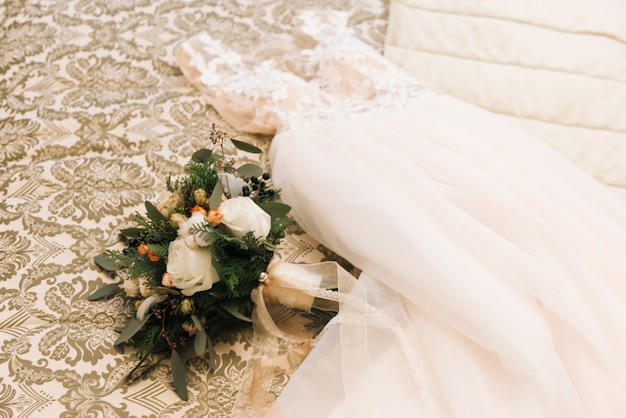 Il bouquet della sposa invernale con rose, cotone, abete rosso si trova accanto all'abito da sposa in una lussuosa camera d'albergo