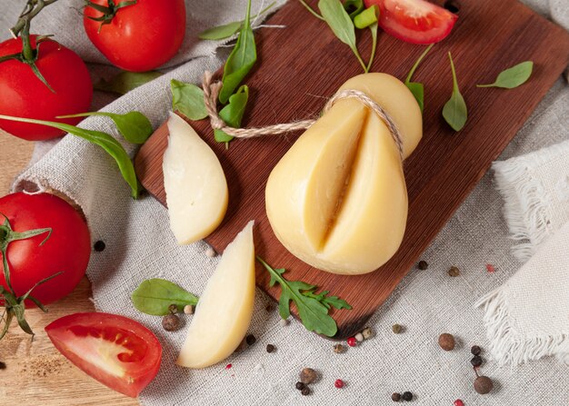 Il caciocavallo è adagiato su una tavola di legno rettangolare. Vengono tagliati due pezzi. Decorate con rucola, spezie e pomodori. Tessuto di lino grigio di sfondo. Vista dall'alto.