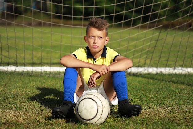 Il calciatore del ragazzo bello in una maglietta gialla si siede vicino alla porta con la palla
