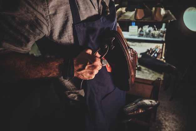 Il calzolaio diligente sta lavorando sulla suola delle scarpe usando uno strumento speciale nel suo posto di lavoro buio.