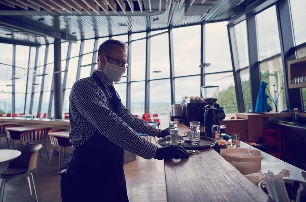 il cameriere con una maschera protettiva medica serve il caffè nel ristorante durante la pandemia di coronavirus che rappresenta un nuovo concetto normale