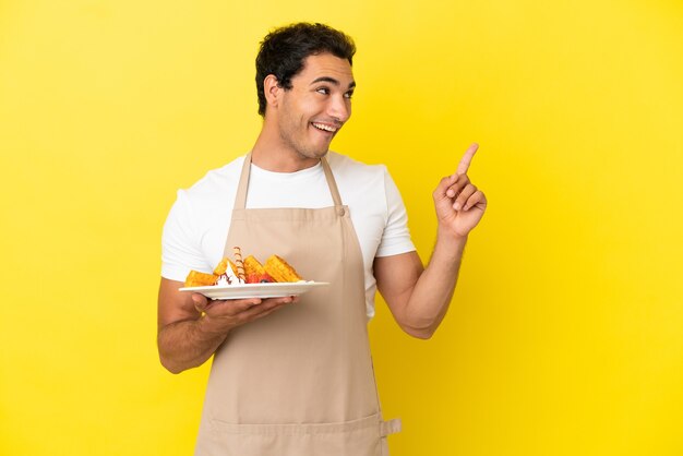 Il cameriere del ristorante che tiene i waffle su uno sfondo giallo isolato con l'intenzione di realizzare la soluzione sollevando un dito verso l'alto