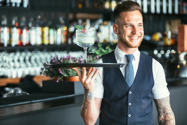 Il cameriere elegante sorridente sta tenendo un vassoio con un cocktail decorato pronto da servire