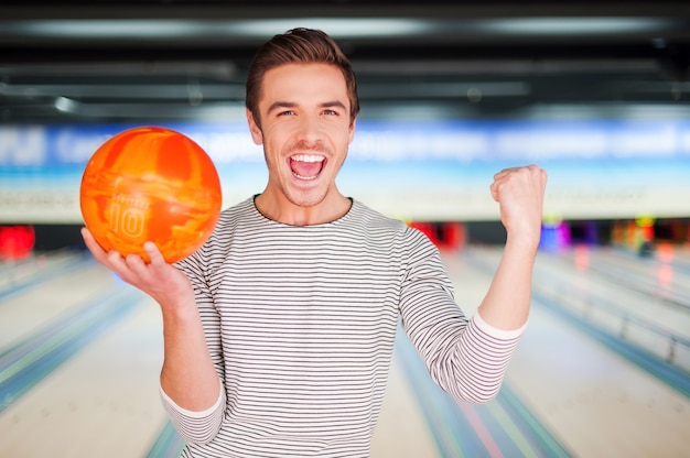 Il campione di bowling. Giovane allegro che tiene una palla da bowling e tiene le braccia alzate mentre si trova in piedi contro le piste da bowling