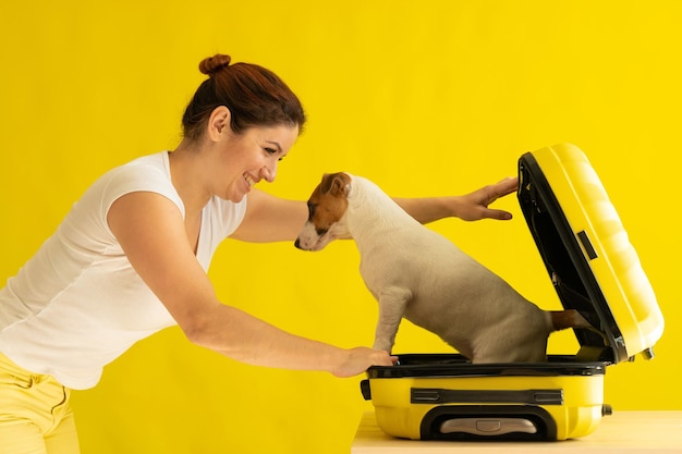 Il cane è seduto in una valigia accanto a una donna che ride su uno sfondo giallo La ragazza sta andando in viaggio con un animale domestico L'amore tra proprietario e cucciolo