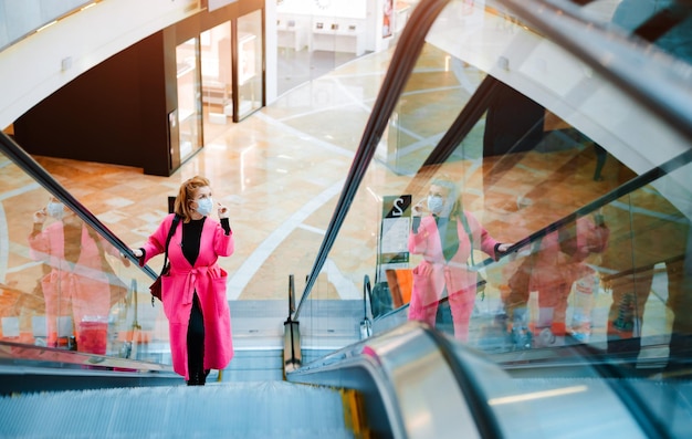 Il cappotto luminoso della bella donna guida la maschera protettiva nera del centro commerciale della scala mobile sul viso contro l'aria inquinata dal virus