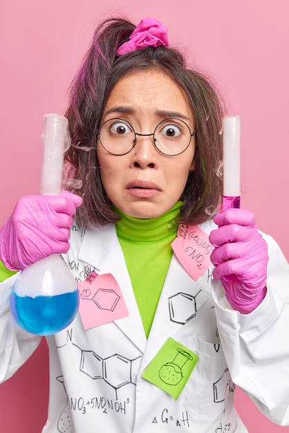 il chimico mescola i reagenti tiene la provetta e il pallone indossa occhiali rotondi il camice medico conduce l'esperimento in pose di laboratorio al coperto sul rosa