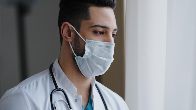 Il chirurgo del terapista uomo arabo premuroso maschio pensieroso indossa una maschera medica uniforme bianca in piedi a