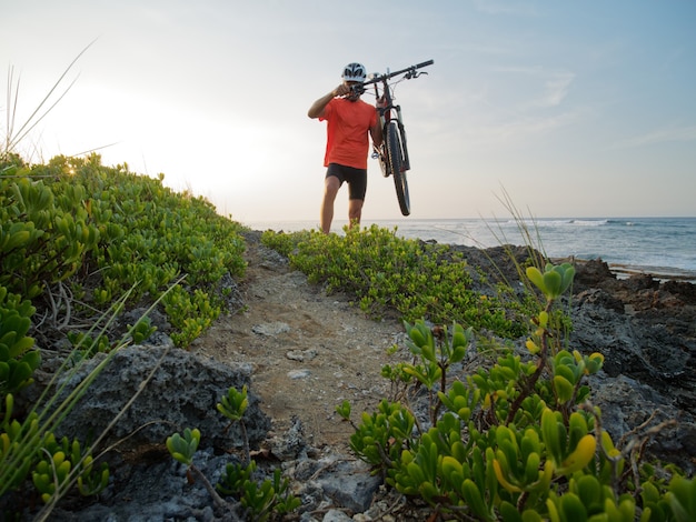 Il ciclista dell'uomo porta una bicicletta nelle sue mani, al litorale dell'oceano. Spiaggia rocciosa.