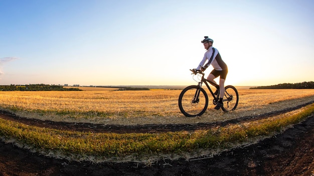 Il ciclista va in bicicletta sulla strada vicino al campo sullo sfondo del sole al tramonto. Sport all'aperto. Uno stile di vita sano.