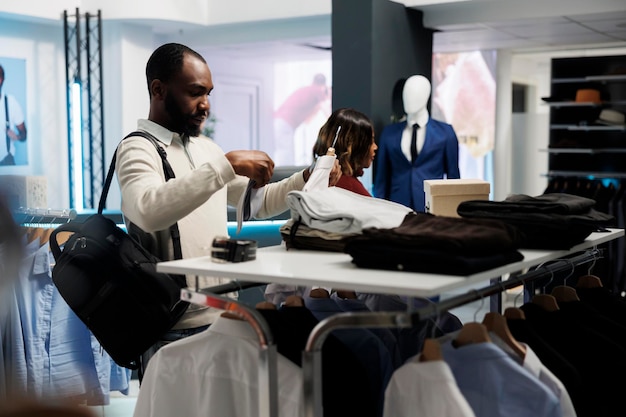Il cliente esamina la cravatta e la camicia durante lo shopping nel negozio di abbigliamento. Uomo afroamericano che sceglie abbigliamento e accessori formali, controllando le dimensioni e lo stile dell'indumento nello showroom